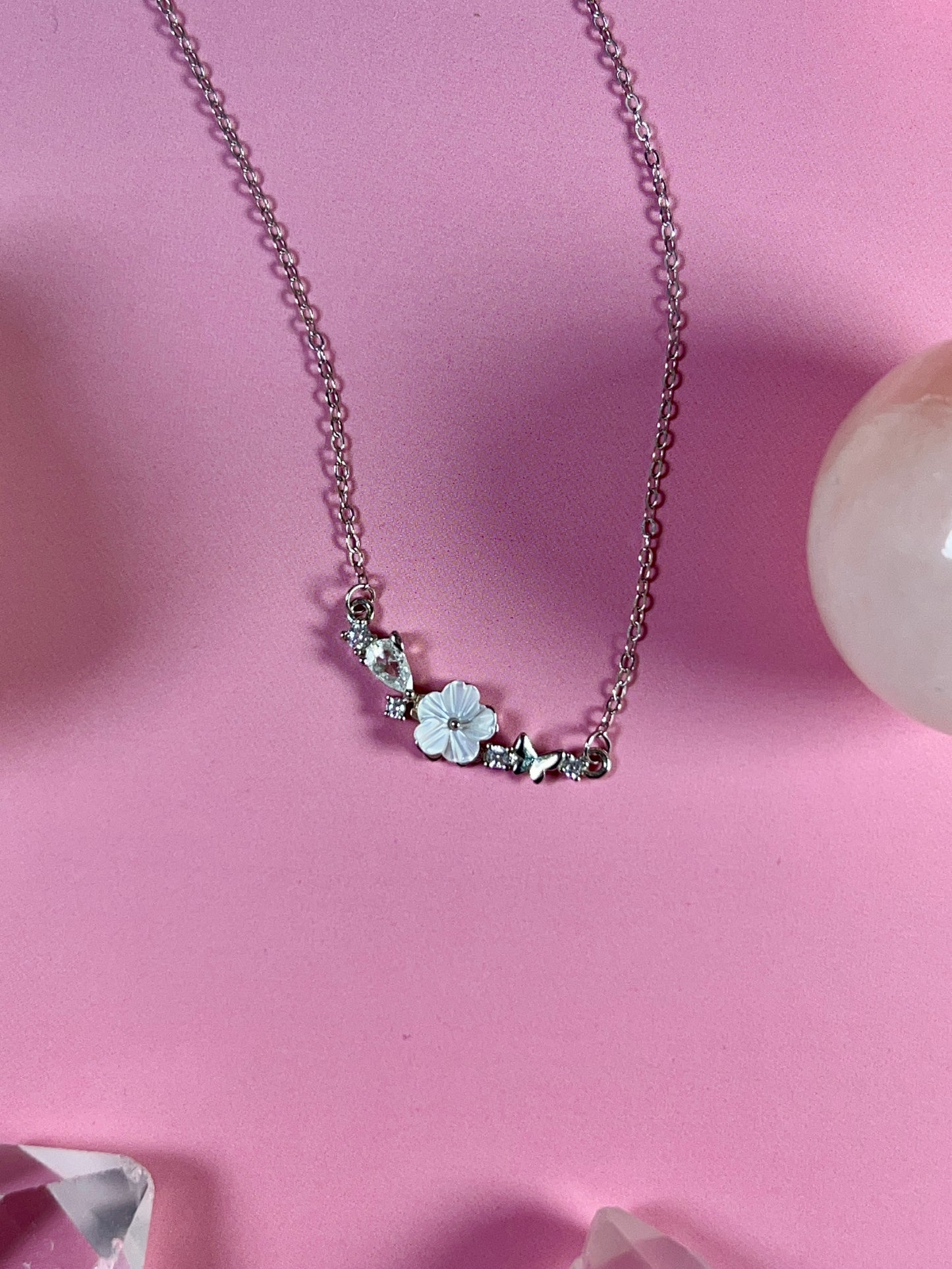 Freya Canada’s I AM FLORA Glass Petals Necklace pictured on a pink background. The necklace features a solid silver butterfly, zircon crystal dew drops and a mother of pearl hand carved flower centre. Ships from Vancouver Bc.