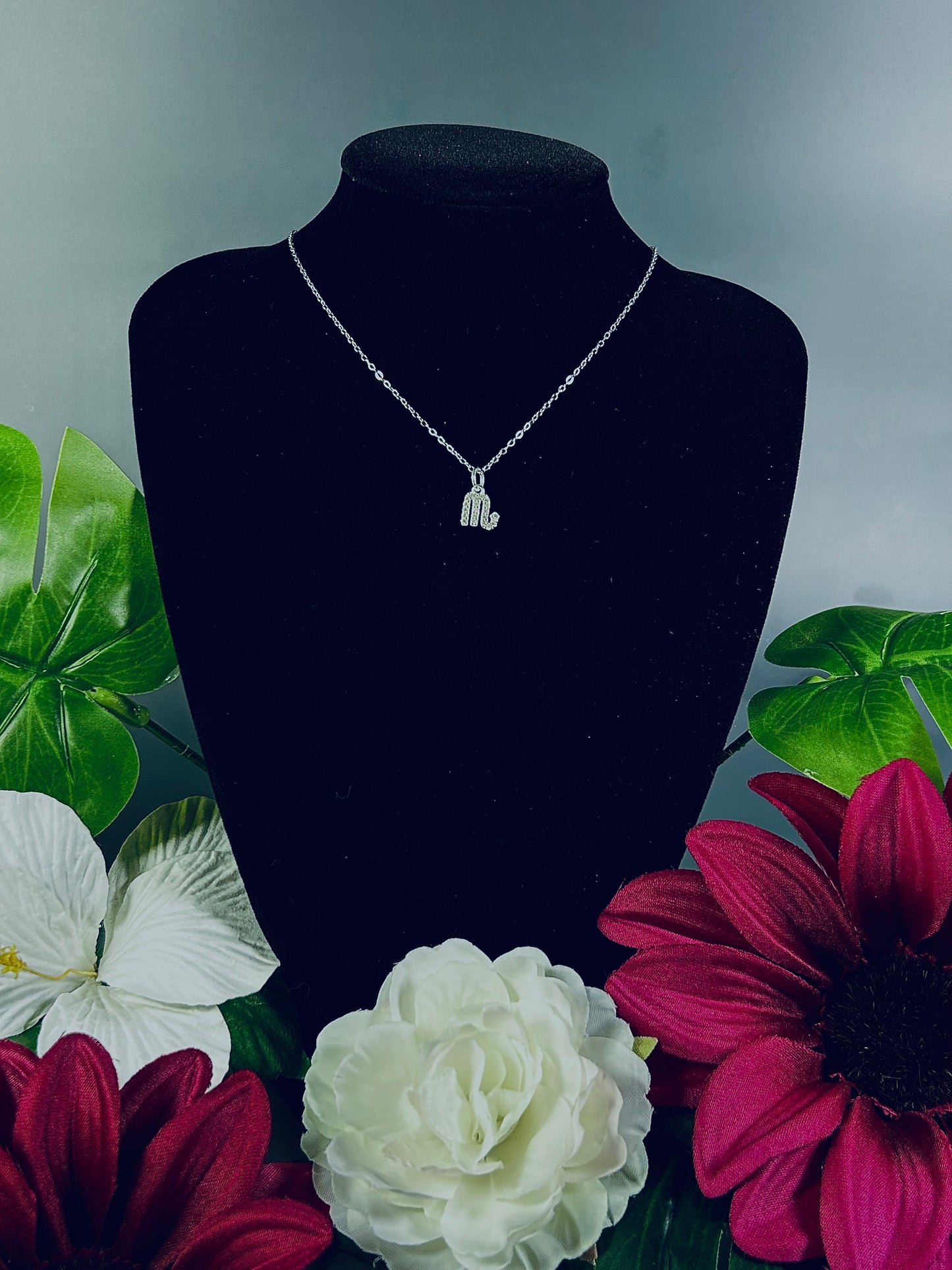 Freya Canada’s Scorpio Goddess Pave Charm Necklace. Pictured with tropical flowers and leaves. Displayed on a black velvet jewelry bust. Ships from Vancouver, B.C.