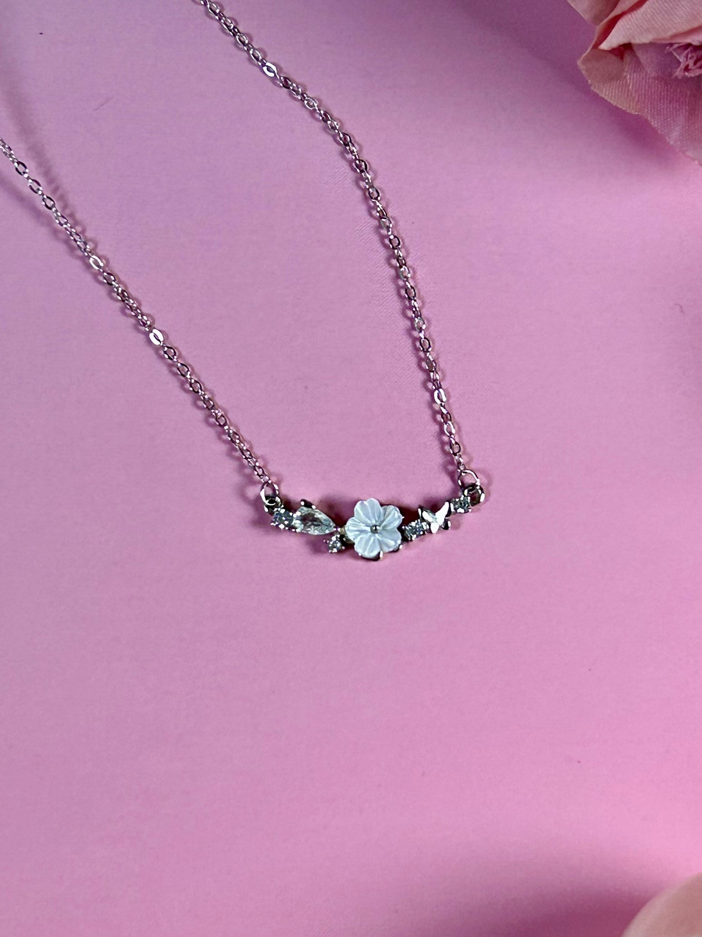 Freya Canada’s I AM FLORA Glass Petals Necklace pictured on a pink satin background. The necklace features a solid silver butterfly, zircon crystal dew drops and a mother of pearl hand carved flower centre.