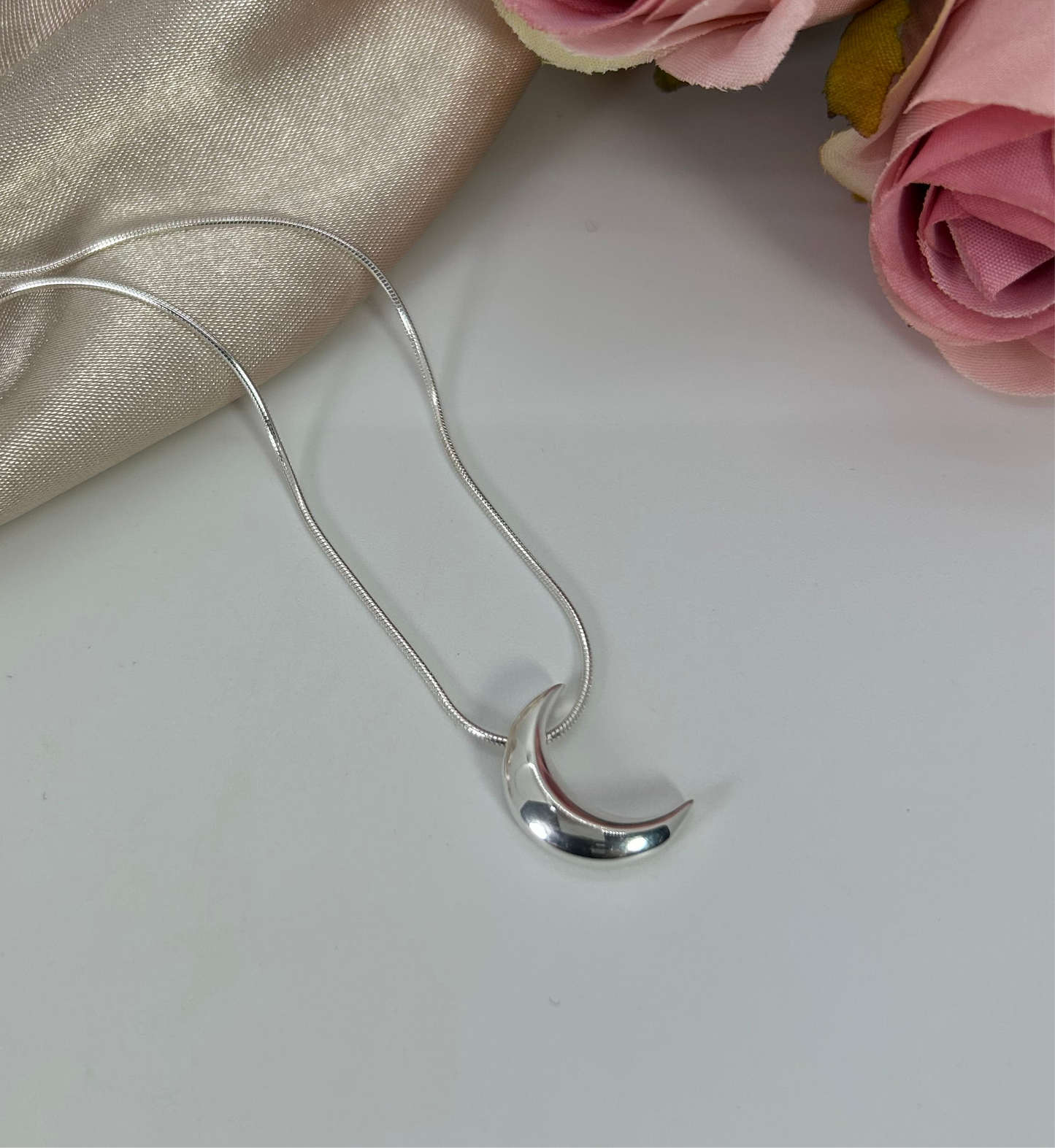 Freya Canada’s I AM SELENE sterling silver luminous crescent moon necklace. Close up, flat lay with pink roses and cream coloured silk fabric elements.