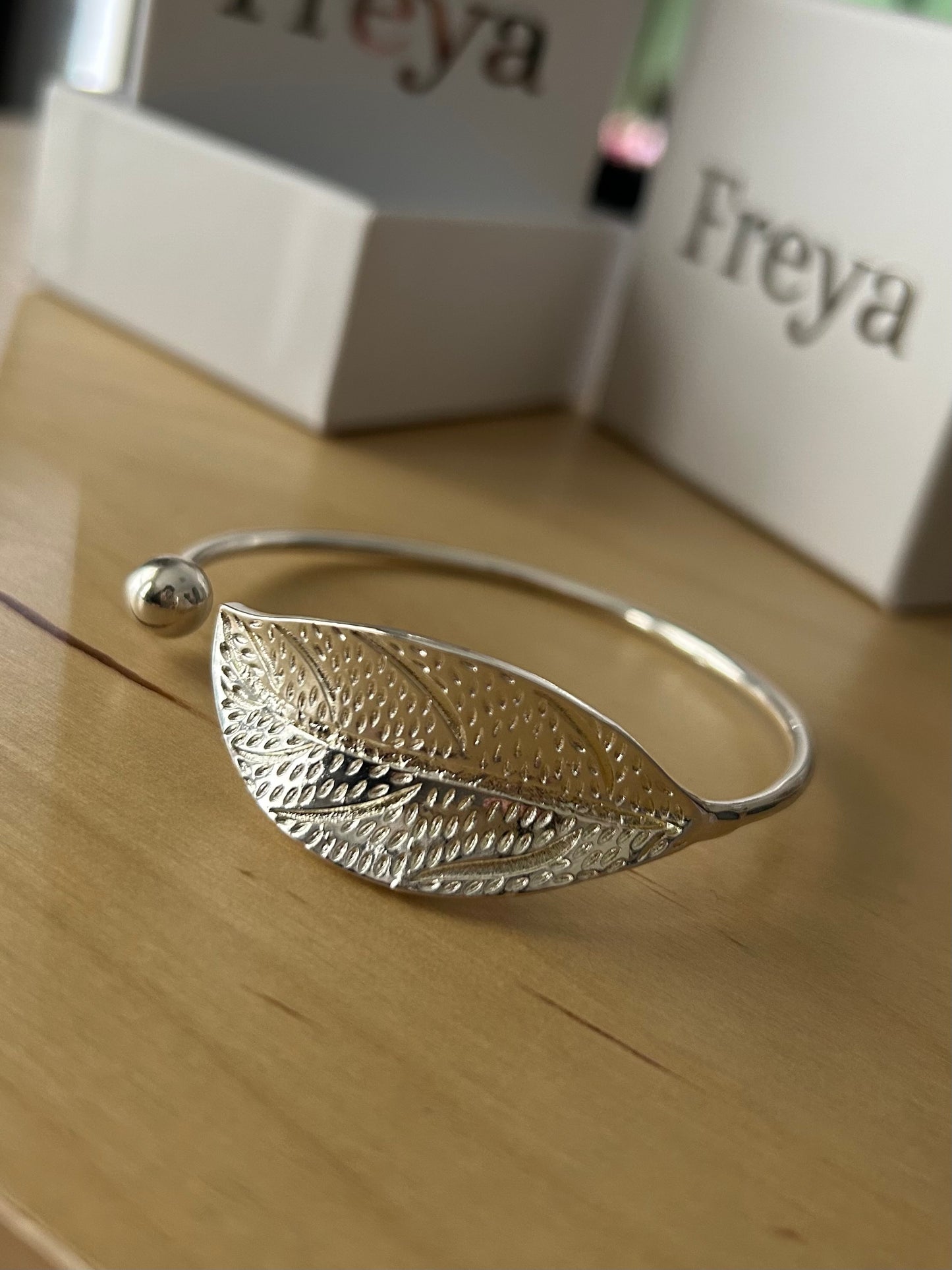 Pictured on a wooden table, the I AM ARTEMIS Wild One Leaf Bangle from Freya Canada. Freya packaging in background. Ships from Vancouver BC.