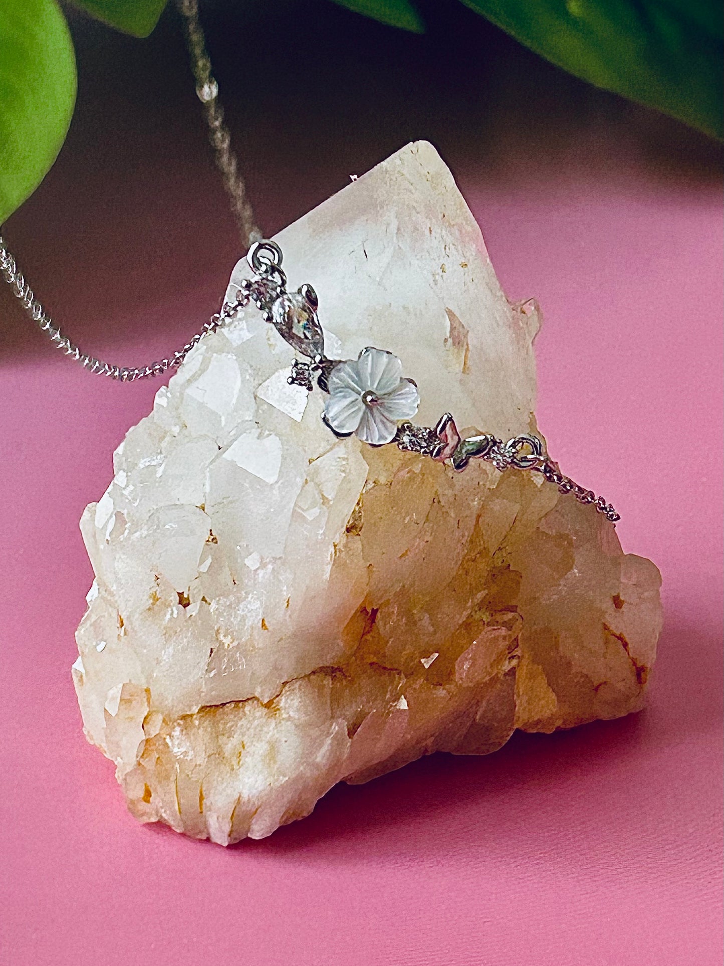 Freya Canada’s I AM FLORA Glass Petals Necklace pictured on a bubblegum pink background with roses. The necklace itself is draped over a quartz crystal cluster and features a butterfly, zircon crystal dew drops and a mother of pearl hand carved flower center