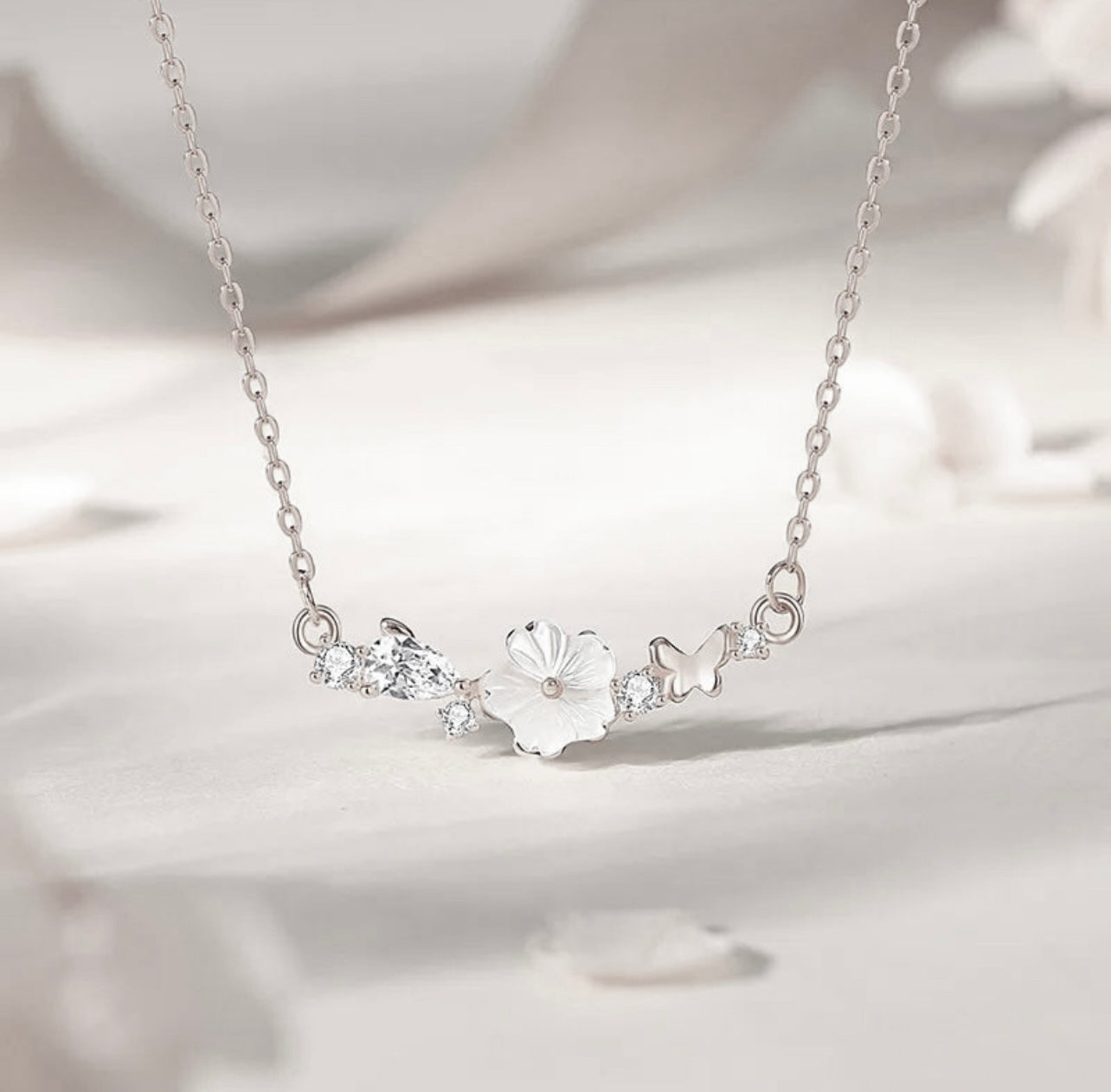 Freya Canada’s I AM FLORA Glass Petals Necklace pictured on a cool white satin background. The necklace features a solid silver butterfly, zircon crystal dew drops and a mother of pearl hand carved flower centre. Ships from Vancouver, British Columbia, Canada