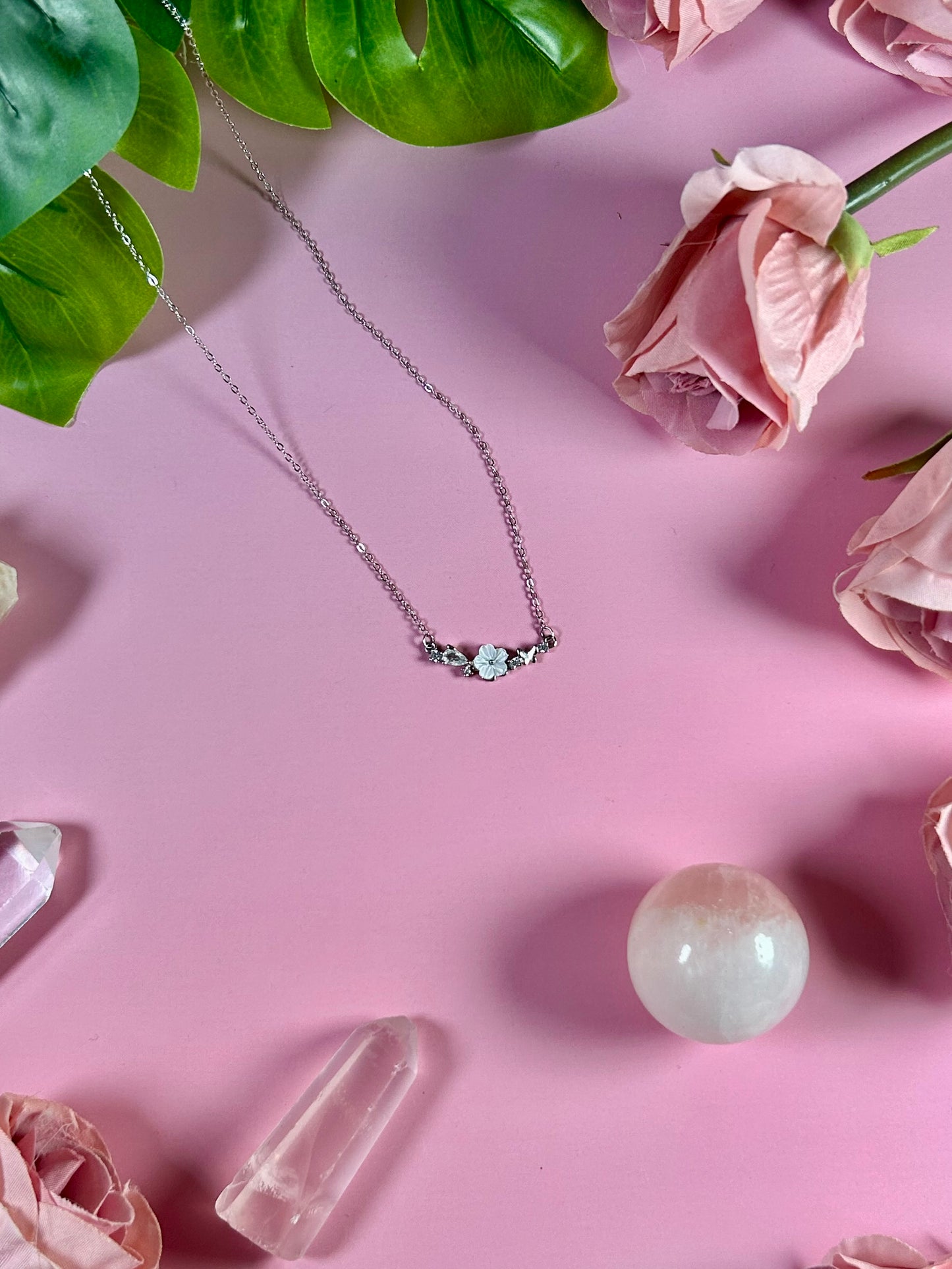 Freya Canada’s I AM FLORA Glass Petals Necklace pictured on a pink satin background with greenery, crystals and roses. The necklace features a solid silver butterfly, zircon crystal dew drops and a mother of pearl hand carved flower centre.