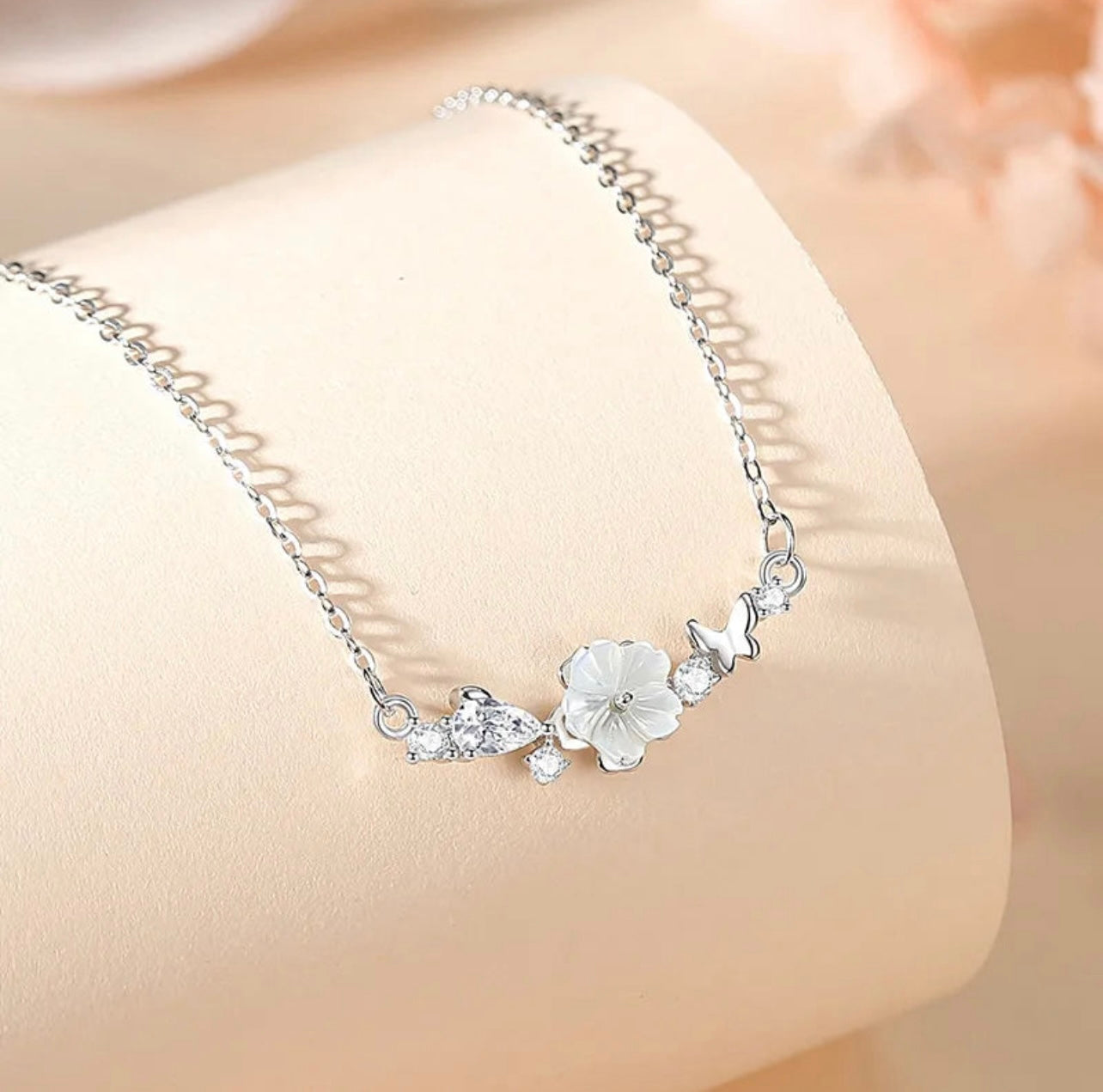 Freya Canada’s I AM FLORA Glass Petals Necklace pictured on a beige satin background. The necklace features a solid silver butterfly, zircon crystal dew drops and a mother of pearl hand carved flower centre