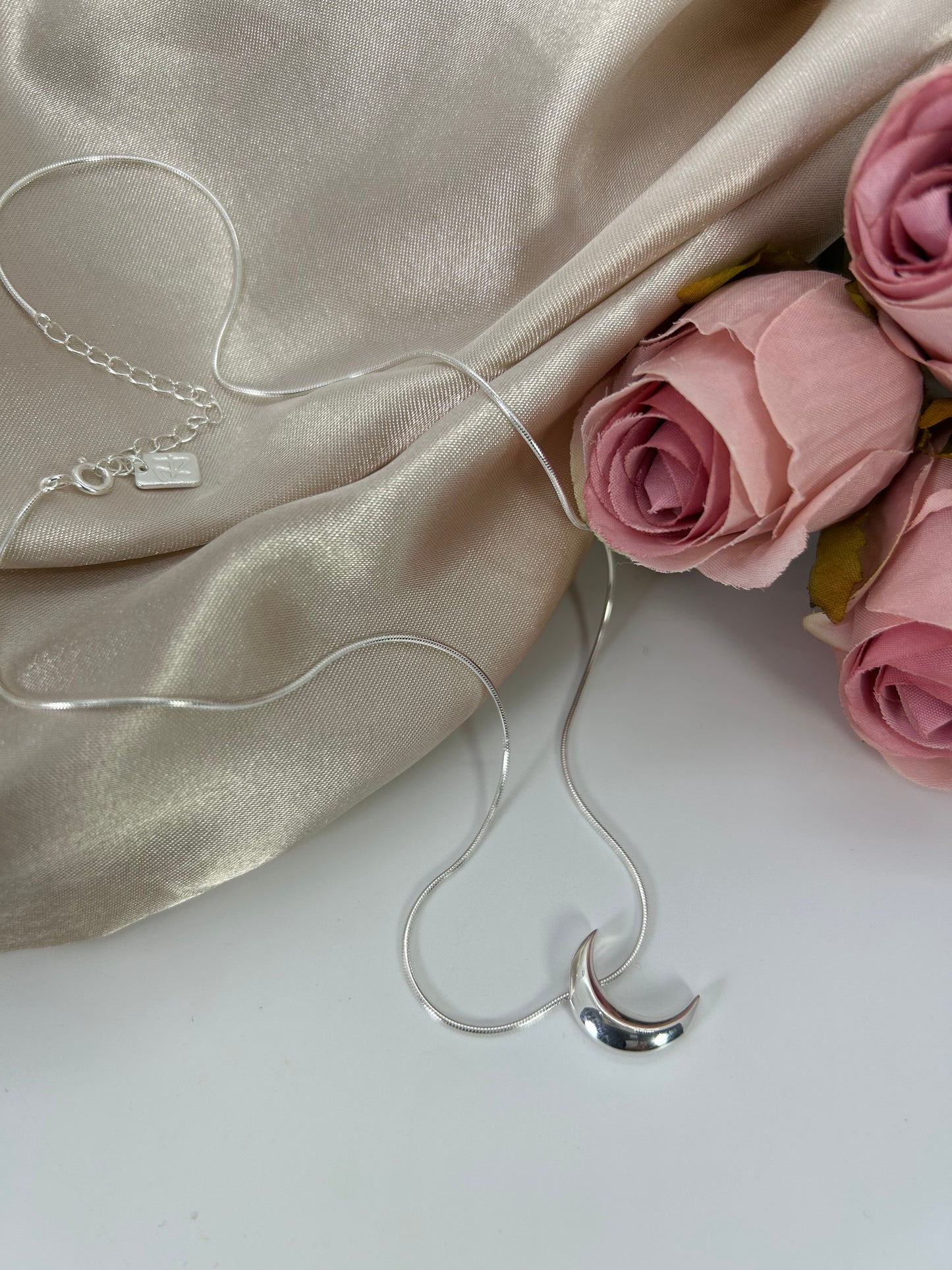 Freya Canada’s I AM SELENE sterling silver luminous crescent moon necklace. Close up, flat lay with pink roses and cream coloured silk fabric elements.