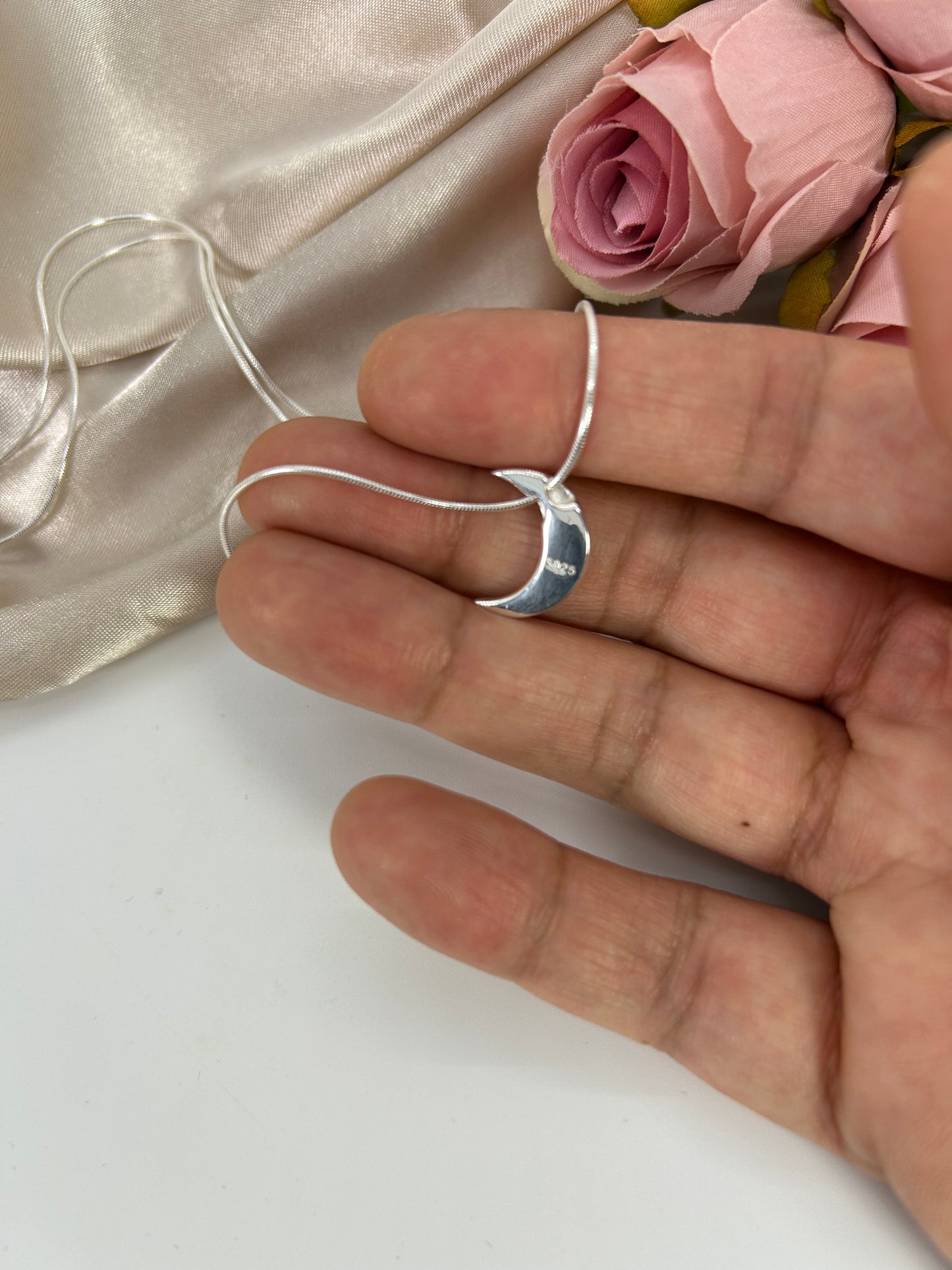 Model is holding Freya Canada’s I AM SELENE sterling silver luminous crescent moon necklace. Showing the back 925 stamp. Close up, flat lay with pink roses and cream coloured silk fabric elements.